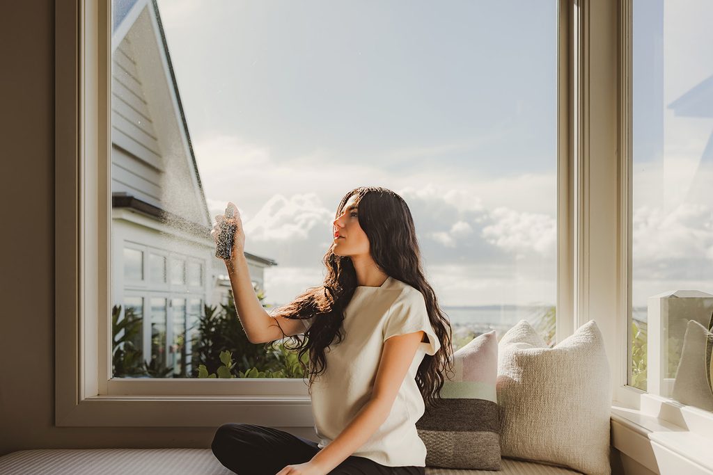 Model applying room spray product by a sunlit window seat with Auckland harbour views in the background"