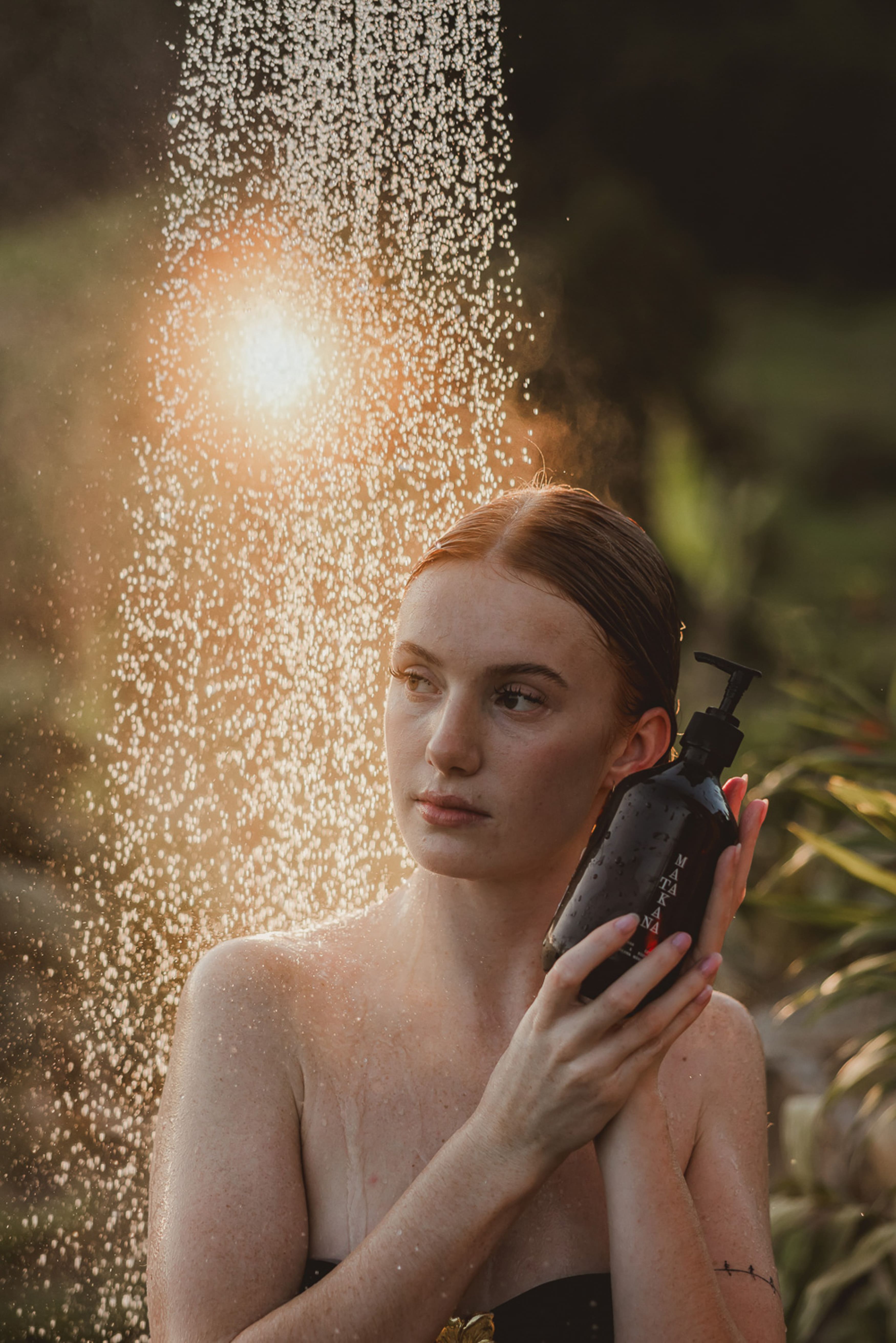 Model holding a hair product bottle in golden backlit water spray, outdoor shower lifestyle shot"
