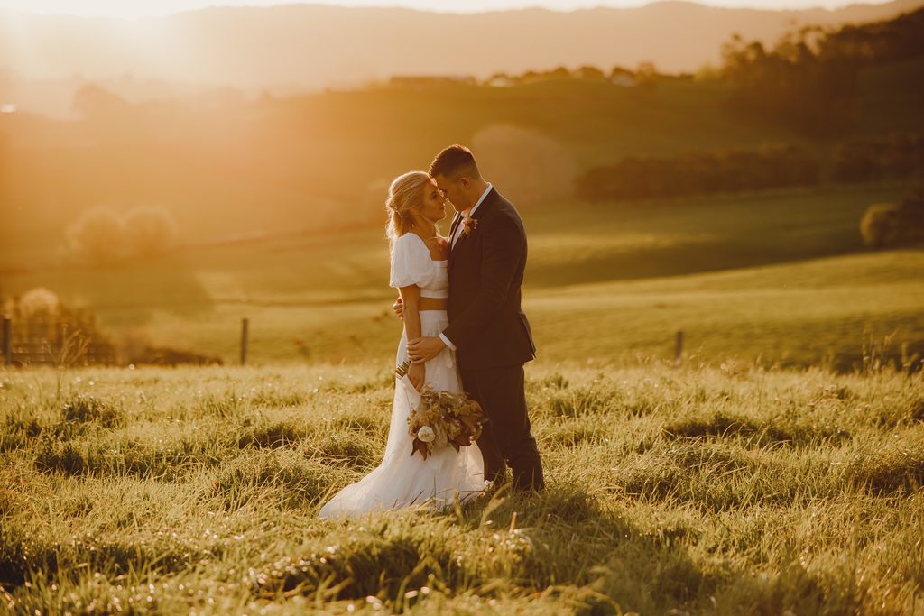 Bride and groom embracing at golden hour in a grassy field, backlit sunset wedding portrait