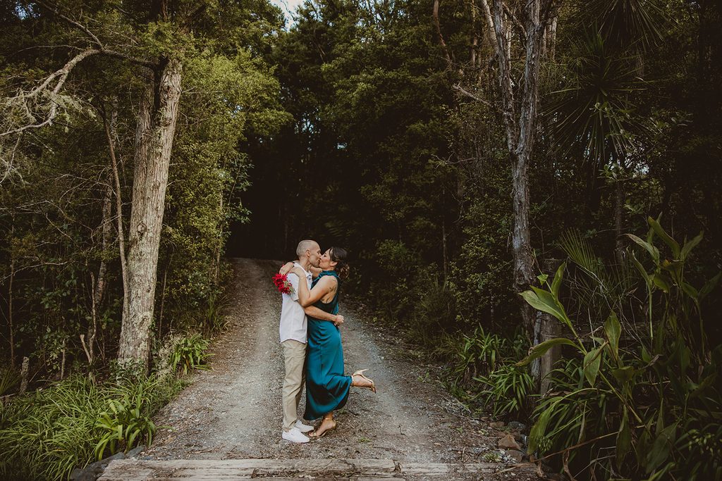 Elopement couple kissing on a bush track at Songbird Retreat, bride mid-kick with red bouquet surrounded by native trees