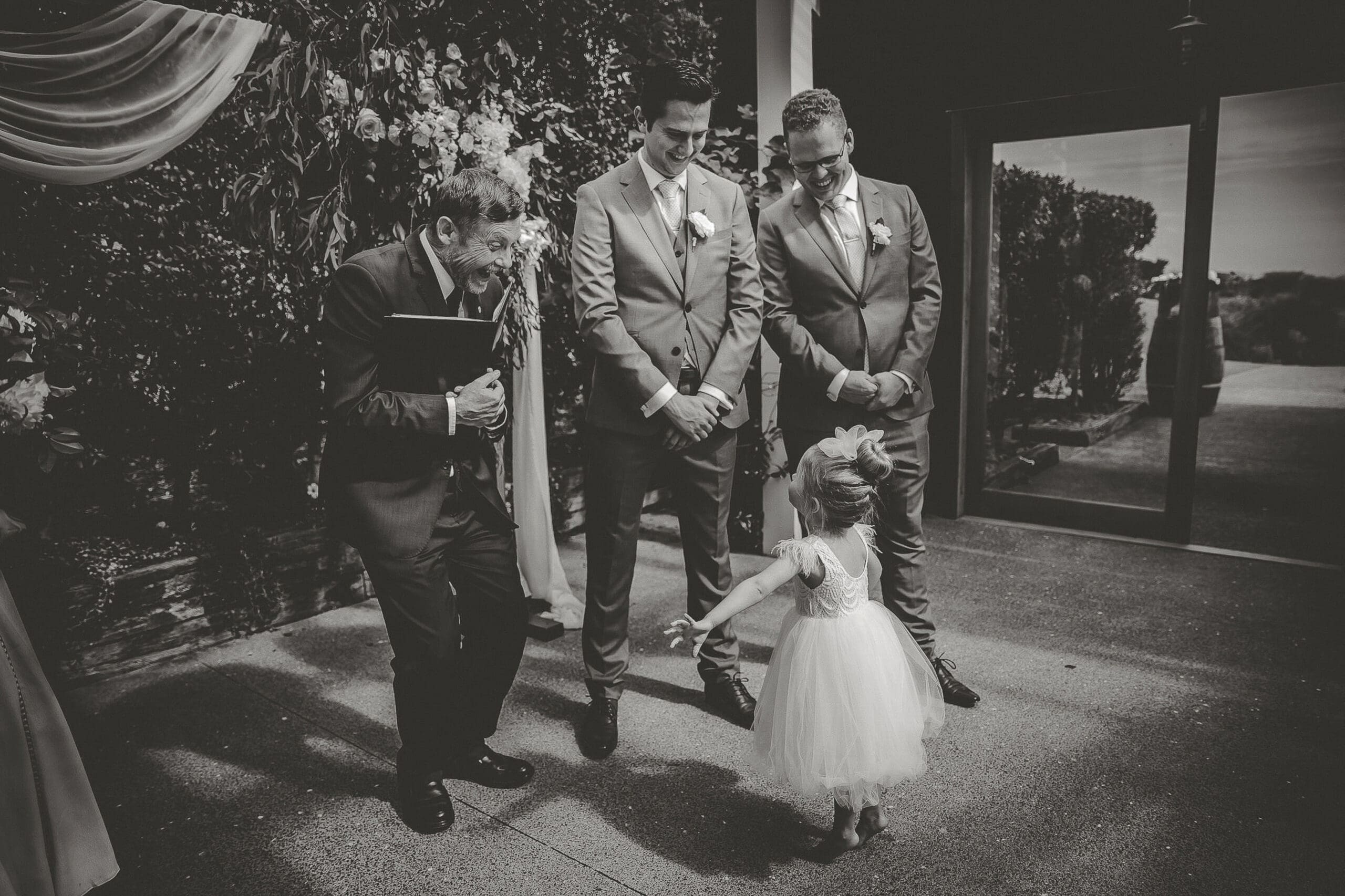 Flower girl showing off her dress to the minister and groomsmen after walking up the aisle, Mudbrick Vineyard wedding, Waiheke Island