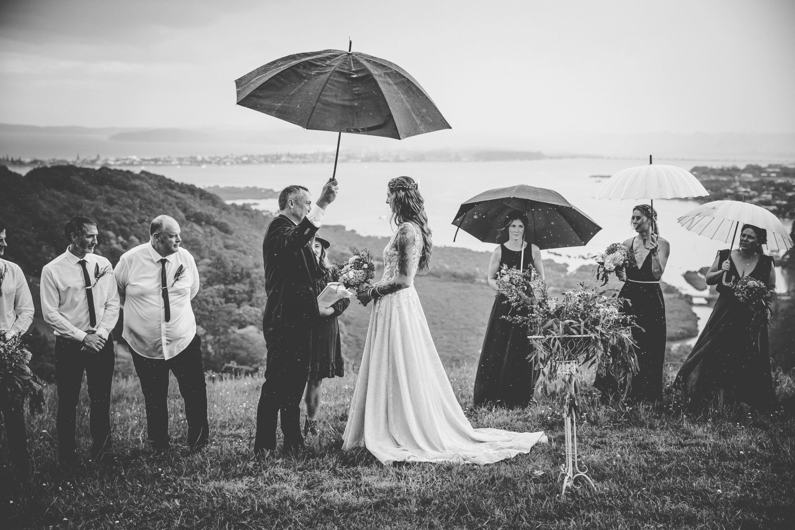 Black and white photo of the bride and groom during their rainy wedding ceremony, surrounded by their bridal party under umbrellas, Matakana coast wedding photographer