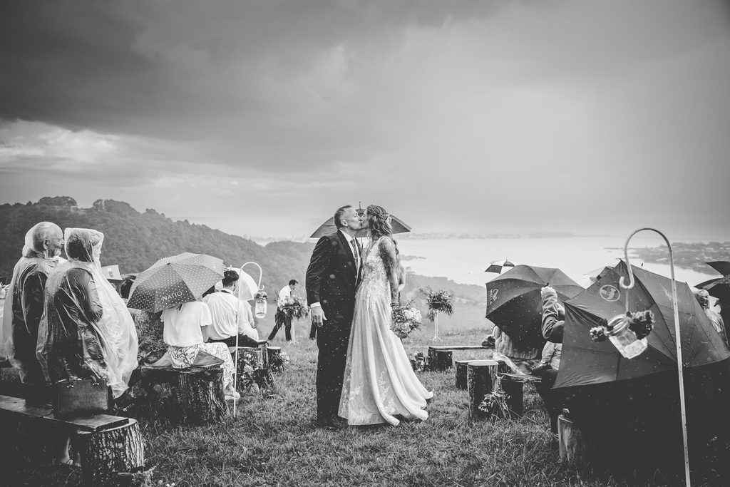 Bride and groom kissing under an umbrella during their rainy wedding ceremony overlooking the coast, Matakana wedding photographer